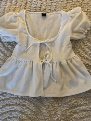 Vit blus med puffärm från Gina Tricot - Supersöt vit blus från Gina Tricot med puffärmar och två knyt framtill. Blusen har en lätt strukturerad yta, djup urringning och volang nedtill. Perfekt för dig som gillar romantisk och trendig stil.