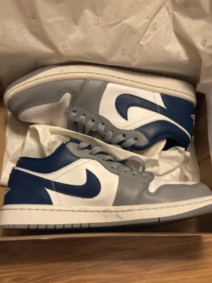 Nike Air Jordan 1 Low blå/grå - Säljer ett par Nike Air Jordan 1 Low sneakers i blått, grått och vitt läder. Klassisk siluett med vit sula, grå snörning och Air Jordan-logga broderad på hälen. Perfekta för dig som gillar streetwear och snygga detaljer.