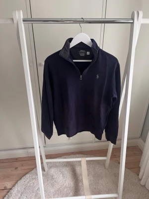 Mörkblå half zip från Polo Ralph Lauren - Mörkblå långärmad tröja från Polo Ralph Lauren med half zip och broderad logga på bröstet. Tröjan har hög krage och är tillverkad i mjukt material, perfekt inför sommaren, passar till allt. Tröjan har blivit lite blekt av solen på framsidan, går säkert att åtgärda, men därav ett lågt pris!Utöver det är tröjan i hyfsat skick med tecken på användning men inga defekter. Skriv vid frågor!
