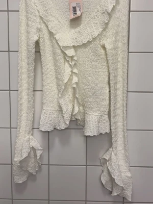 Vit volangblus med lång ärm - Supersöt vit blus med volangdetaljer längs krage, ärmslut och nederkant. Blusen har en smockad struktur och långa ärmar med extra volang vid handlederna. Perfekt för dig som gillar romantisk och trendig stil.