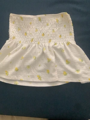 Vit blommig bandeautopp Gina Tricot - Supersöt bandeautopp från Gina Tricot i vitt med små gula blommor. Toppen har smock upptill och en volangig nederdel som ger en luftig känsla. Perfekt för varma dagar och enkel att styla med jeans eller kjol.