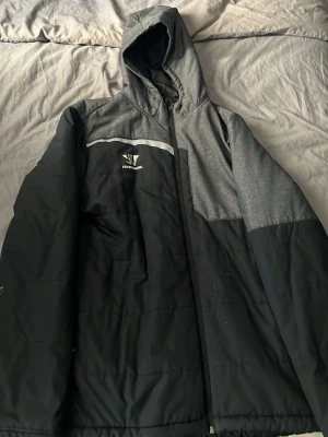 Svart och grå pufferjacka med huva - Snygg svart och grå pufferjacka från Streamlight med huva och dragkedja. Jackan har en kontrasterande grå axelparti och huva, samt en vit detalj över bröstet. Perfekt för kalla vinterdagar och har en sportig look.