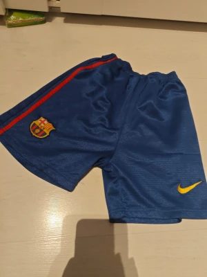 Blåa FC Barcelona shorts Nike - Snygga blåa FC Barcelona shorts från Nike med klubbmärke på ena benet och gul Nike-logga på det andra. Shortsen har en röd rand längs sidan och är gjorda i ett lätt och ventilerande material, perfekt för fotbollsträning eller match.