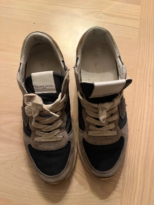 Philippe Model sneakers i svart och beige - Snygga sneakers från Philippe Model med svart textil och beige mockadetaljer. Skorna har rund tå, platt sula och vita snören. Insidan är fodrad med ljus skinn. Slitna under annars är sicket helt okej. 