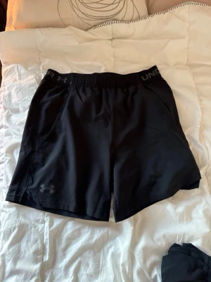 Svarta träningsshorts från Under Armour - Säljer ett par svarta träningsshorts från Under Armour med elastisk midja och logga på både midjebandet och benet. Shortsen är tillverkade i ett lätt och snabbtorkande syntetmaterial, perfekta för intensiva träningspass. Storlek M