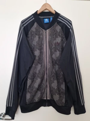 Svart Adidas herr track jacket med mönster - Snygg Adidas Originals träningsjacka i mörkgrå/svart med unikt mönstrat framparti. Klassiska tre ränder längs ärmarna och dragkedja hela vägen framtill. Storlek 2XL. Säljs pga stor viktnedgång.  I riktigt fint skick – inga fläckar eller skador. Enda lilla anmärkningen är att det grå partiet runt kragen har börjat flagna/ljusna lite, men det syns inte vid användning.   Detta är ett plagg som är riktigt svårt att få tag på idag – perfekt för samlare och adidas-fans