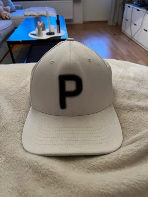 Vit keps med svart P-bokstav - Snygg vit keps med böjd skärm från Pumal. Klassisk sexpanelsdesign med ventilationshål och justerbar baksida. Perfekt accessoar för en clean och sportig look. Materialet känns som bomull eller polyester.