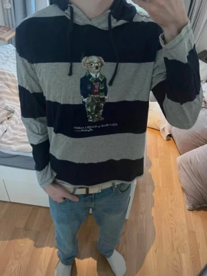 Randig hoodie Polo Bear Ralph Lauren - Säljer en grå och marinblå randig hoodie från Ralph Lauren med den ikoniska Polo Bear-figuren på bröstet. Hoodien har huva med dragsko och är tillverkad i mjukt bomullsmaterial. Perfekt för dig som vill ha en chill och stilren look.