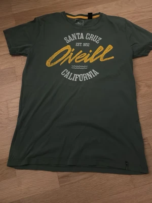 Grön O'Neill t-shirt Santa Cruz - Snygg grön t-shirt från O'Neill med tryck i vitt och gult framtill där det står 'Santa Cruz California'. Slim fit-modell i mjuk ekologisk bomull. Perfekt för en avslappnad och cool stil.