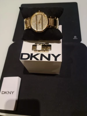 Guldig DKNY klocka med stenar - Snygg guldig klocka från DKNY med glittriga stenar på urtavlan och armbandet. Klockan har en rund form och metallarmband med DKNY-logga på spännet. Perfekt accessoar för att lyfta din stil.