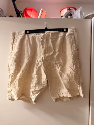 Beiga linneshorts med snörning - Säljer ett par beiga shorts i linne med avslappnad passform och snörning i midjan. Shortsen har fickor på sidorna och en bakficka. Perfekta för varma dagar och ger en chill vibe till din outfit.