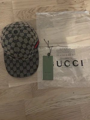 Gucci beige keps med GG-mönster - Snygg beige keps från Gucci med klassiskt GG-monogram över hela kepsen. Kepsen har en böjd skärm och en röd-blå rand på sidan som ger extra lyxig känsla. Tillverkad i canvasmaterial och levereras med originalpåse och tagg.