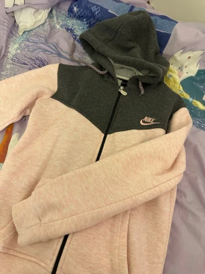 Nike hoodie i rosa och mörkgrå - Nike hoodie med dragkedja i en snygg färgkombination av ljusrosa och mörkgrå. Hoodien har huva med dragsko, ribbade muddar och fickor framtill. Nike-logga broderad på bröstet i rosa. Perfekt för en chill och sportig look.