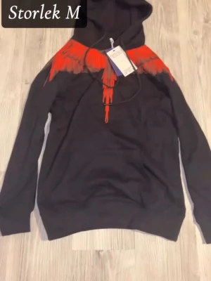 Svartröd hoodie - Säljer en svart hoodie från Marcelo Burlon med ikoniskt rött vingmönster över axlar och bröst. Hoodien har huva med dragsko och en klassisk känguruficka framtill. Materialet är mjukt och bekvämt, perfekt för en cool streetwear-look.