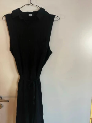 Svart ärmlös skjortklänning med knappar - Svart ärmlös skjortklänning med krage och knappar framtill. Klänningen har ett knytband i midjan och en bröstficka. Perfekt för en stilren och enkel look. Rök och djurfritt hem. Pruta gärna 🌸