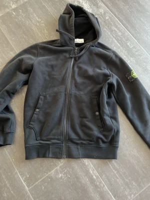 Svart hoodie från Stone Island - Svart hoodie från Stone Island med dragkedja framtill, två fickor med knappstängning och klassisk logopatch på ärmen. Tillverkad i mjukt bomullsmaterial och har en stor huva med dragsko. Perfekt för en avslappnad och stilren look.