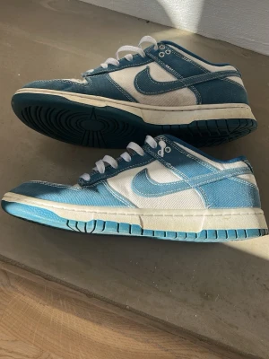Nike Dunk Low blå/vit denim - Säljer ett par Nike Dunk Low sneakers i blått och vitt med denimliknande material. Skorna har vita snören, vit sula och klassisk Nike Swoosh på sidan. Tåboxen har små blå stjärnor som detalj. 