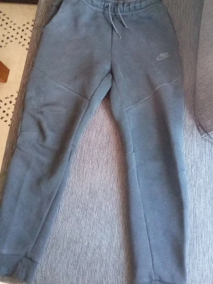 Nike mjukisbyxor - Säljer ett par mörkgrå mjukisbyxor från Nike med dragsko i midjan och diskret Nike-logga på vänster lår. Byxorna har en slim fit passform och är tillverkade i mjukt, tjockt material som känns riktigt skönt mot huden. Perfekta för chill eller träning.