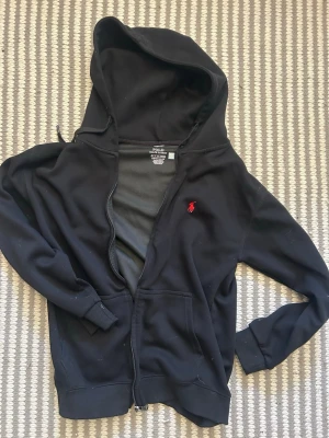Svart hoodie från Polo Ralph Lauren - Svart hoodie med dragkedja från Polo Ralph Lauren. Klassisk design med huva och dragsko, samt röd broderad logga på bröstet. Tillverkad i mjukt bomullsmaterial och har två fickor framtill. Perfekt för en avslappnad och stilren look.