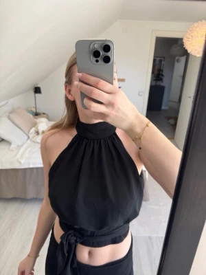 Svart halterneck topp med knyt - Trendig svart halterneck topp med hög krage och bred knytning i midjan. Toppen är ärmlös och har en croppad passform som framhäver midjan. Perfekt för dig som vill ha en stilren och modern look.