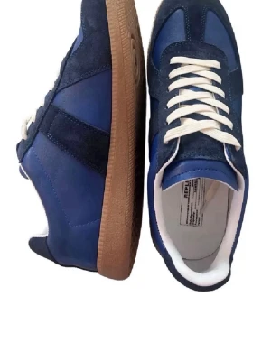 Blå sneakers från Maison Margiela - Snygga blå sneakers från Maison Margiela med ovandel i mocka och läder, vita skosnören och klassisk gummisula i brunt. Skorna har en låg silhuett och stilrena detaljer som passar till många outfits.
