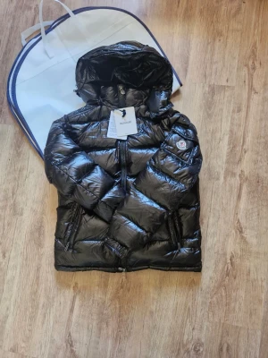 Moncler Jacka storlek 4 - Snygg svart pufferjacka från Moncler med glansig finish och stor huva. Jackan har två dragkedjefickor fram, tydlig Moncler-logga på ärmen och quiltad design. Perfekt för kalla dagar och riktigt varm tack vare dunfyllning. Storlek 4 eller XL eller L. Snabb leberans.