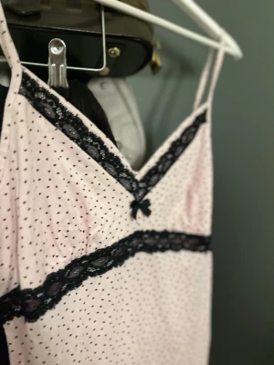 Rosa nattlinne med spetsdetaljer - Sött nattlinne i ljusrosa med små svarta prickar och tunna axelband. Dekorerat med svart spets längs ringningen och midjan samt en liten rosett framtill. Materialet ser mjukt och bekvämt ut, perfekt för mysiga kvällar.
