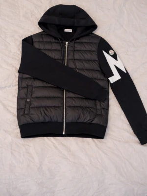 Svart Moncler cardiganjacka med huva - Snygg svart cardiganjacka från Moncler med vadderad kropp och mjuka ärmar. Jackan har en stor vit Moncler-logga på ena ärmen, dragkedja framtill och två sidofickor. Huva i sweatshirtmaterial ger en sportig look. Perfekt för dig som vill ha något trendigt och bekvämt.