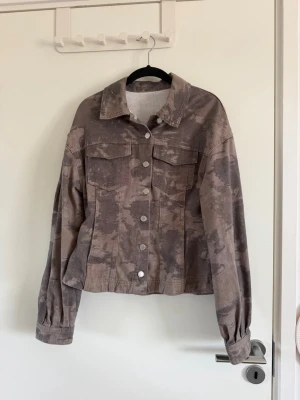 Camo-mönstrad jeansjacka i brunt - Snygg jeansjacka med camo-mönster i olika bruna och grå toner. Klassisk modell med krage, två bröstfickor med lock och silverfärgade knappar framtill. Jackan har en avslappnad passform och långa ärmar med knapp vid ärmslut.