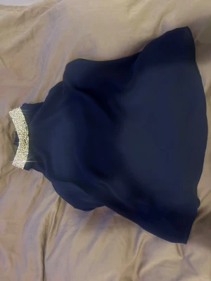 Mörkblå ärmlös blus med paljetter - Snygg mörkblå blus i ett lätt, flytande tyg med en glittrig paljettkant runt halsen. Blusen är ärmlös och har en avslappnad passform som ger en elegant look. Perfekt att styla med jeans eller kjol för en trendig outfit.