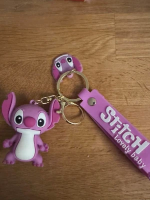 Lila Stitch nyckelring med figur - Nyckelring med en lila Stitch-figur i mjukt gummi och matchande band med texten 'Stitch Lovely Baby'. Söt design med stora öron och vita detaljer, perfekt att hänga på väskan eller nycklarna.