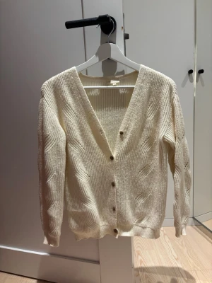 Krämvit kabelstickad kofta - Krämvit kabelstickad kofta med v-ringning och ribbade muddar. Koftan har dekorativa kabelstickade detaljer längs ärmar och framstycke samt knappar framtill. Storleken är 146/152 men passar mig som är S