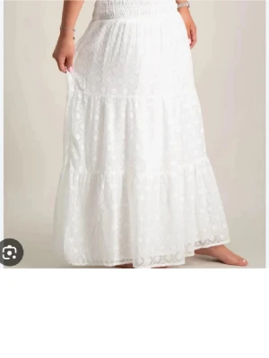 Vit långkjol med spetsdetaljer - Superfin vit långkjol med volangdetaljer och diskret mönster. Kjolen har en smockad midja som ger en bekväm passform och ett luftigt fall. Perfekt för varma dagar och ger en romantisk vibe till din outfit.