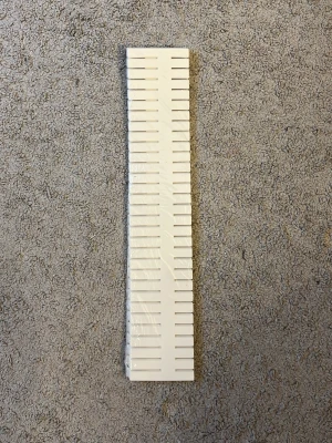 Vit plaststicka med spår - Lång, rektangulär vit plaststicka med jämnt fördelade spår längs båda sidor. Perfekt för att organisera eller hålla ordning på småsaker som kablar eller trådar. Enkel och stilren design som passar i många olika sammanhang.