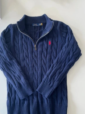 Marinblå kabelstickad tröja Polo Ralph Lauren - Kabelstickad marinblå tröja från Polo Ralph Lauren med halv dragkedja och hög krage. Tröjan har det klassiska röda Polo-logot broderat på bröstet och ribbade muddar vid ärmslut och nederkant. Perfekt för lager på lager och riktigt snygg till jeans.