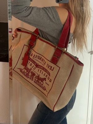 Levi's beige/röd handväska - Jättecool vintage handväska från Levi's i beige med röda detaljer & Levis tryck framtill. Väskan har röda handtag i läder, metallspännen och Levi's motivet. Perfekt storlek för att få plats med allt du behöver, har olika fack inuti och passar till det mesta🥰💕Unisex passform. 