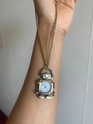 Halsband med robotklocka i metall - Unikt halsband med en robotformad hängare i antikguldfärgad metall. Robotens kropp har en inbyggd klocka med vit urtavla och svarta siffror. Kedjan är tunn och matchar robotens färg. Perfekt accessoar för dig som gillar coola och annorlunda smycken.helt funkar