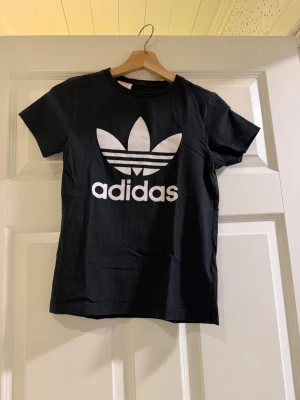 Svart Adidas t-shirt med logga - Snygg svart t-shirt från Adidas med den klassiska vita trefoil-loggan och texten på bröstet. T-shirten har rund halsringning och korta ärmar. Tillverkad i mjuk bomull för skön känsla och enkel stil.