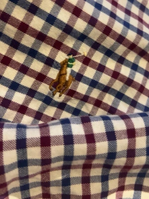 Rutig skjorta från Ralph Lauren - Snygg rutig skjorta från Ralph Lauren med klassiskt broderat logotyp på bröstet. Skjortan har blå, vinröda och vita rutor, knappar framtill och lång ärm. Perfekt för en stilren och avslappnad look.