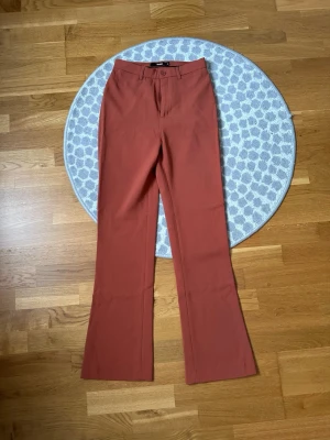 Rostfärgade kostymbyxor från Bik Bok - Snygga kostymbyxor från Bik Bok i en trendig rostfärg. Byxorna har raka ben med lätt utsvängning, klassisk midja med hällor och stängs med knapp och dragkedja. Perfekta för dig som vill ha en stilren look med lite edge.