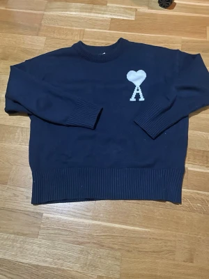 Mörkblå stickad tröja från Ami - Mörkblå stickad tröja från Ami Alexandre Mattiussi med rund halsringning och ribbade muddar. På bröstet finns ett vitt hjärta med bokstaven A. Perfekt för en stilren och trendig look. Komm med bud vid snabb affär 