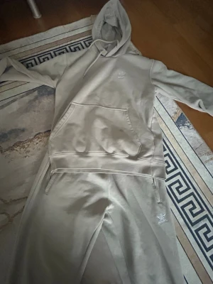 Beige Adidas loungewear set - Säljer ett beige loungewear set från Adidas med matchande hoodie och byxor. Hoodien har magficka och dragsko i huvan, medan byxorna har resår i midjan och sidofickor. Båda plaggen har Adidas logga broderad framtill. Perfekt för chill dagar hemma.