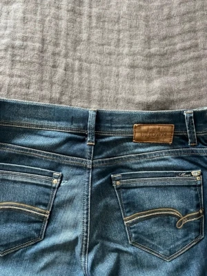 Vintage blå bootcut jeans från Uptown  - Säljer ett par blå bootcut jeansmed snygga detaljer på bakfickorna och klassisk läderpatch i midjan. Jeansen har en lätt tvättad look och är tillverkade i denim med kontrastsömmar. Perfekta för dig som gillar en tidlös och avslappnad stil. I storlek 38 men passar även 36 för en mer baggy look. 
