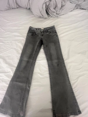 Gråa bootcut jeans - Säljer ett par gråa bootcut jeans med klassisk femficksdesign och slitna detaljer. Jeansen har hög midja och vida ben, perfekta för en avslappnad och trendig look. Materialet är mjukt denim i bomull. Passar dig som gillar retrostil.