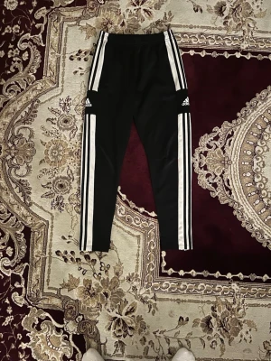 Svarta Adidas träningsbyxor - Svarta träningsbyxor från Adidas med klassiska vita ränder längs sidorna och logga på låret. Byxorna har smal passform, resår i midjan och är tillverkade i ett stretchigt material som passar perfekt för sport och träning.