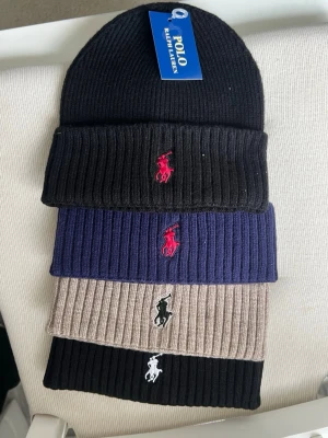 Ribbstickade mössor från Polo Ralph Lauren - Fyra ribbstickade mössor från Polo Ralph Lauren i svart, marinblått och beige. Varje mössa har den klassiska broderade loggan framtill i kontrasterande färg. Perfekt accessoar för att hålla stilen under kalla dagar. 200kr/st 