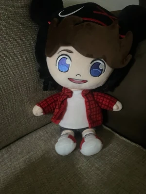 Kreekcraft plushie - Söt mjukisdocka med stora blå ögon, brunt hår och rödsvart rutig skjorta över en vit t-shirt. Dockan har svarta shorts och vita skor med röda detaljer. Perfekt som prydnad eller leksak.