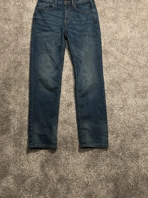Blå raka jeans med fem fickor - Säljer ett par klassiska blå jeans med rak passform och fem fickor. Jeansen har normal midja och subtila slitningar framtill för en avslappnad look. Tillverkade i denim med snygga kontrastsömmar. Perfekta till vardagsstilen!