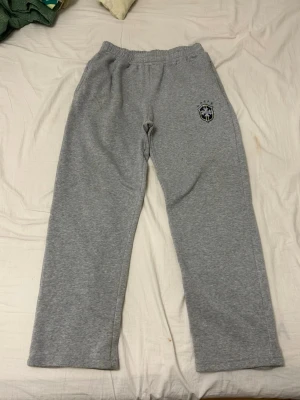 Grå mjukisbyxor med Saint-logga - Säljer ett par grå mjukisbyxor från Saint med broderad logga på vänster lår. Byxorna har resår i midjan och raka ben. Tillverkade i mjukt sweatshirtmaterial som är perfekt för chill dagar. Klassisk och enkel design med sportig touch.