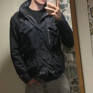 Mörkblå Field Jacket från Ralph Lauren - Mörkblå field jacket från Ralph Lauren med flera stora fickor, dragkedja och snörning i midjan. Jackan har hög krage och huvan går att stoppa in.